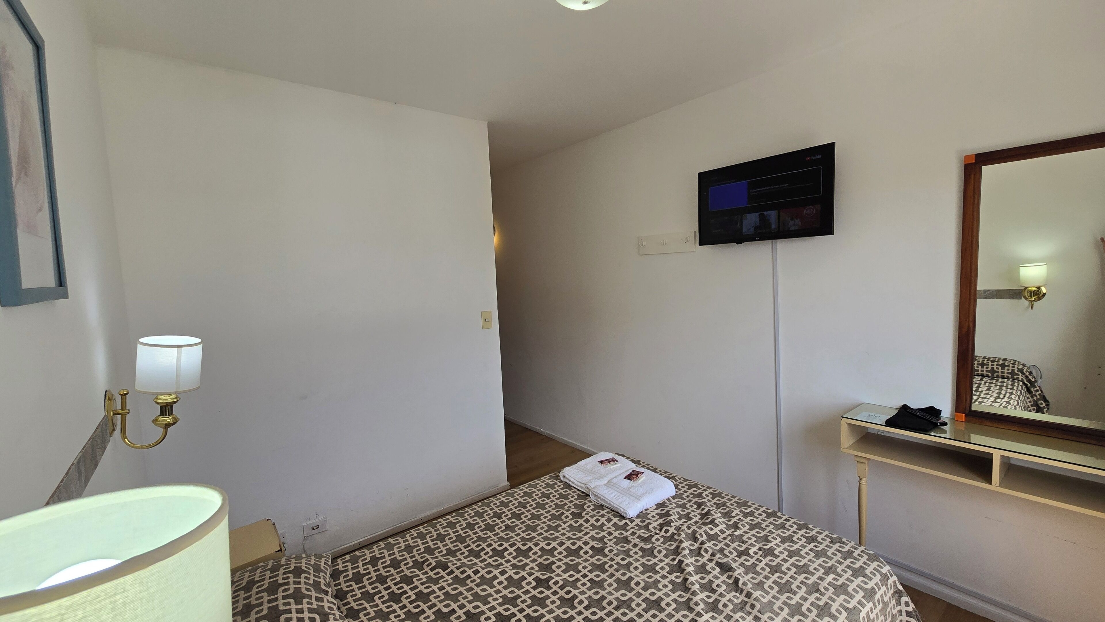 A hotel room with a bed, a bedside table with a lamp, a TV mounted on the wall, and a mirror.