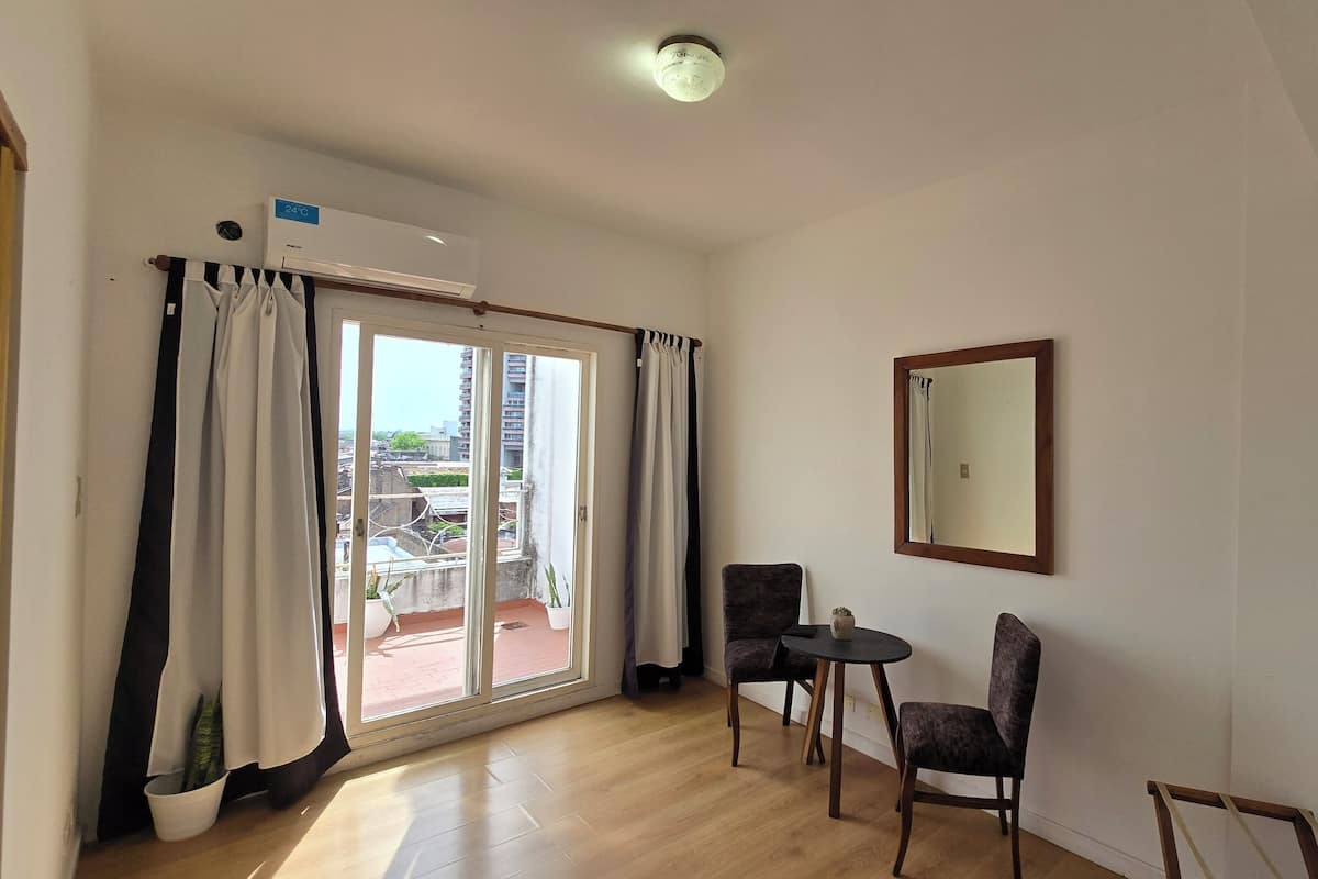 Ein Zimmer mit einer Schiebetür aus Glas, die zu einem Außenbereich führt, einer Decken-Klimaanlage, zwei Stühlen, einem kleinen Tisch und einem Spiegel an der Wand.