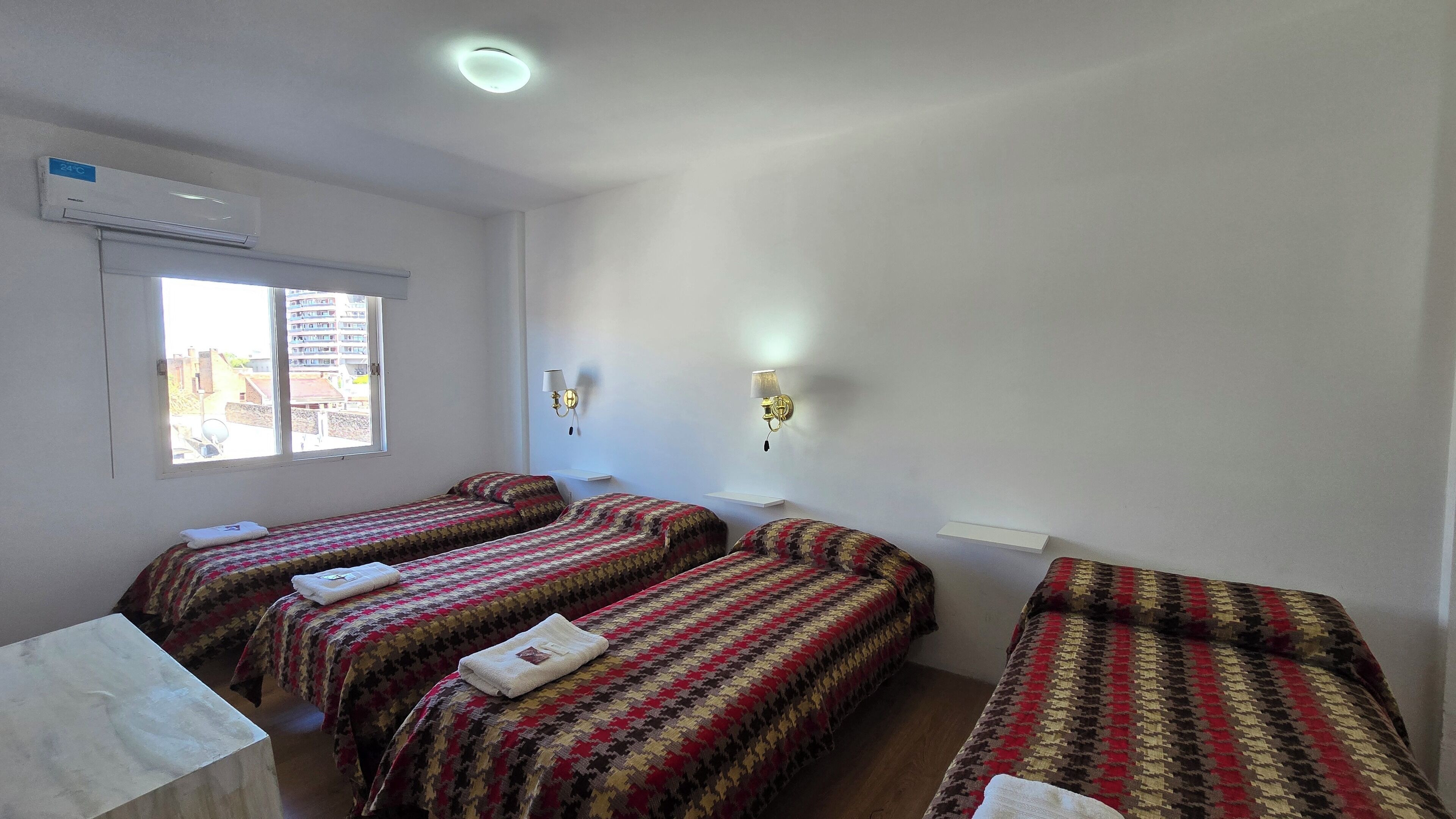 A room with three beds, a window with a view of buildings, an air conditioning unit, and a wall-mounted lamp.