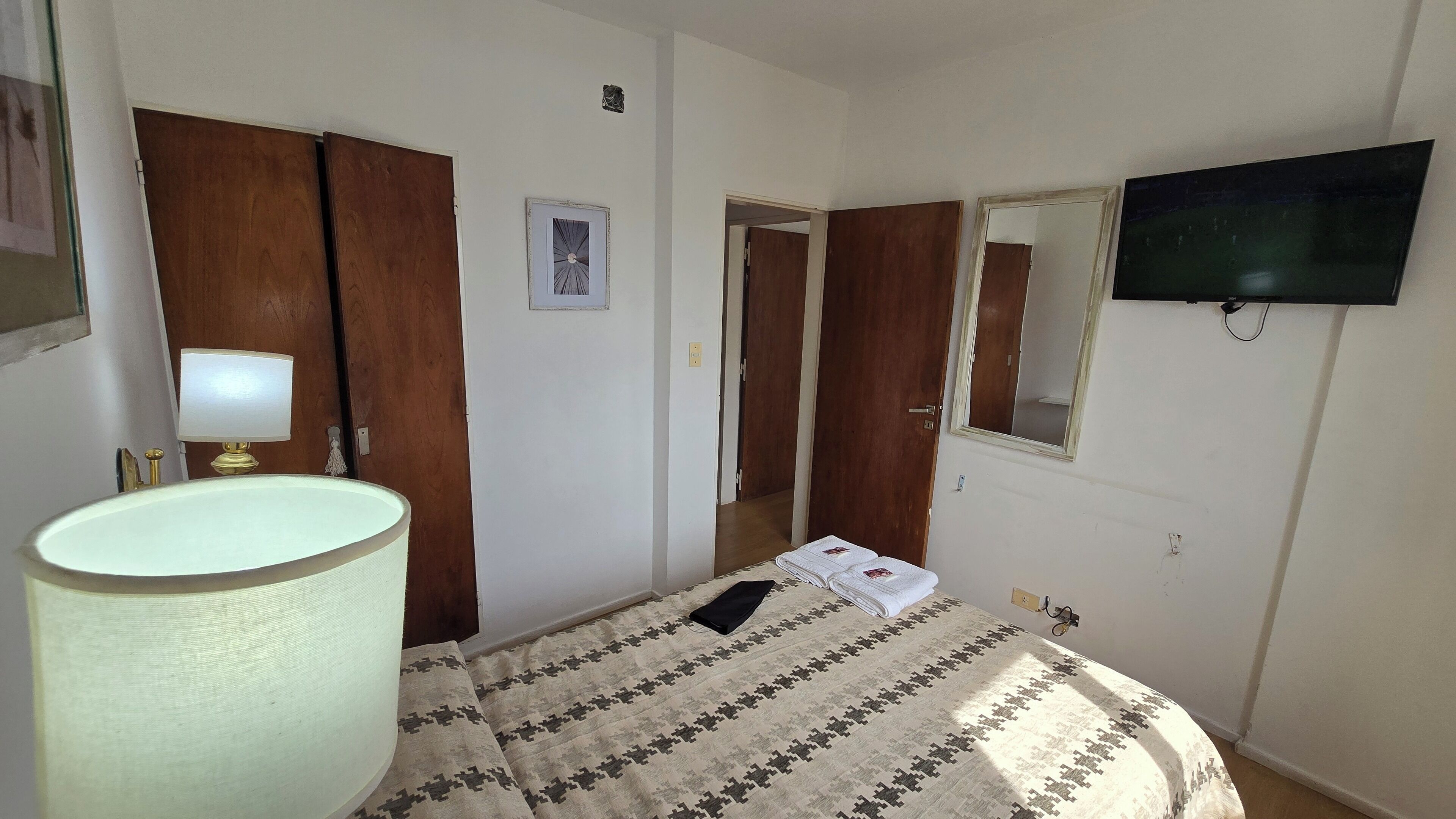 Een hotelkamer met een bed, een bedlampje, een aan de muur bevestigde televisie en een deur naar een andere kamer.