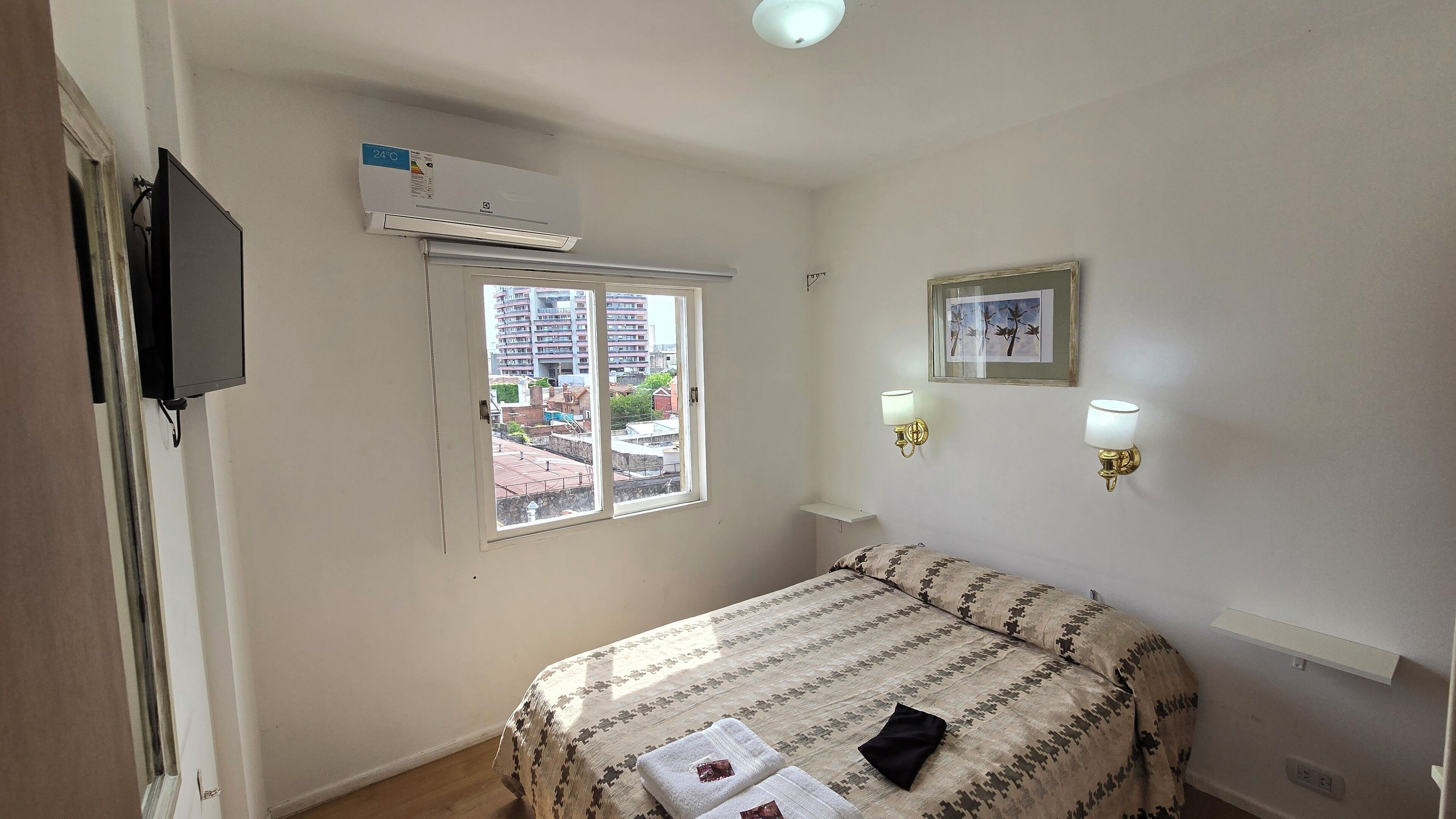 A hotel room with a bed, a window with a view of buildings, a mounted TV, and wall-mounted lights.