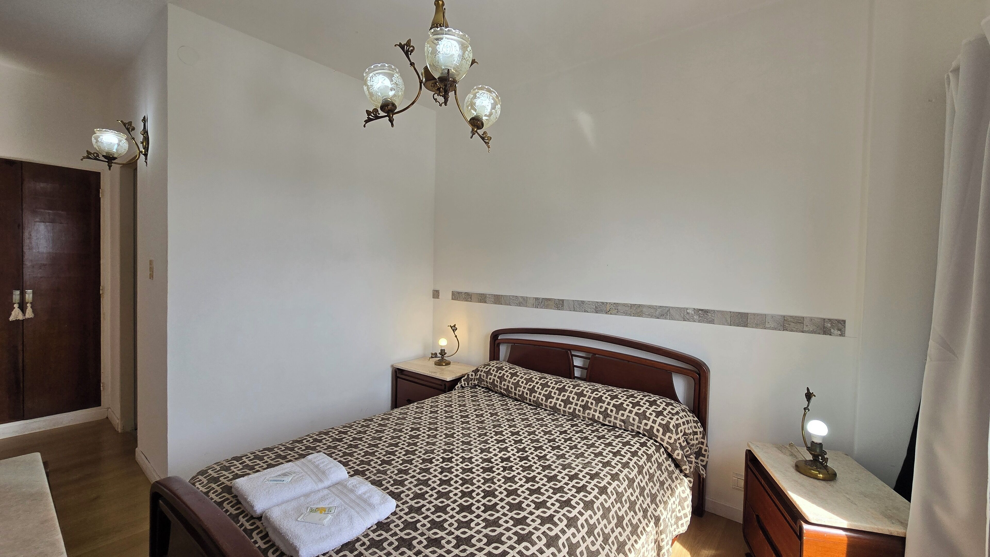 A bedroom with a patterned bedspread, a wooden wardrobe, a chandelier, and bedside lamps.