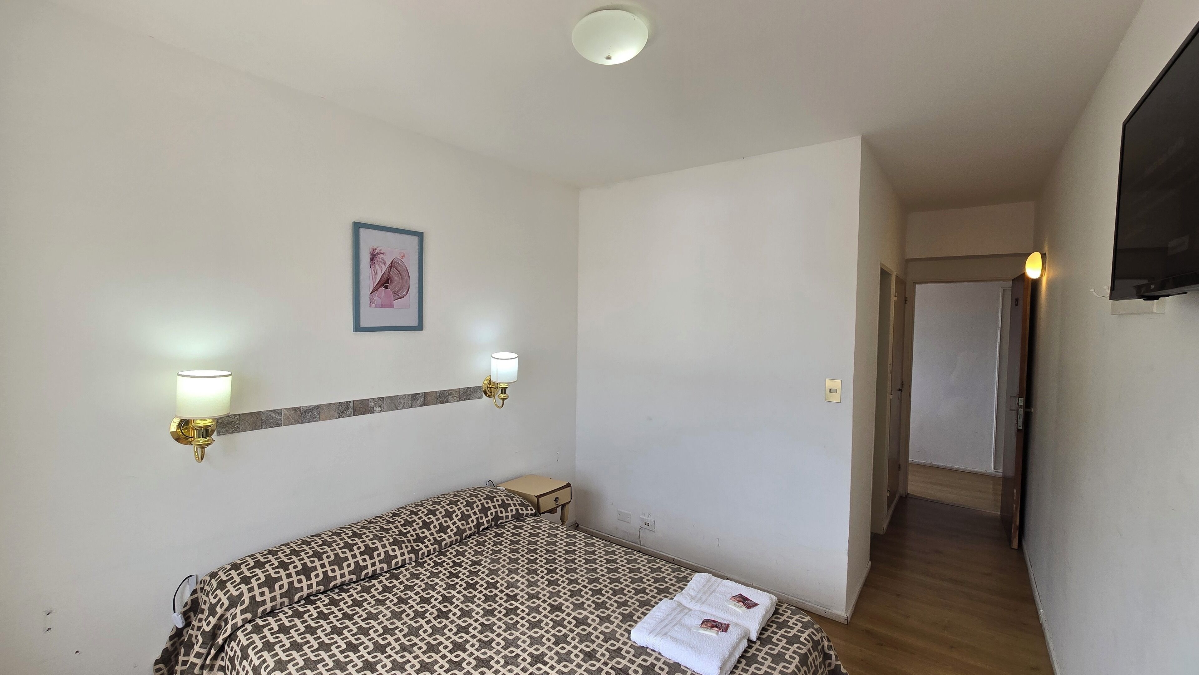A small, clean room with a bed, two wall-mounted lights, a framed picture, and a door leading to another room.