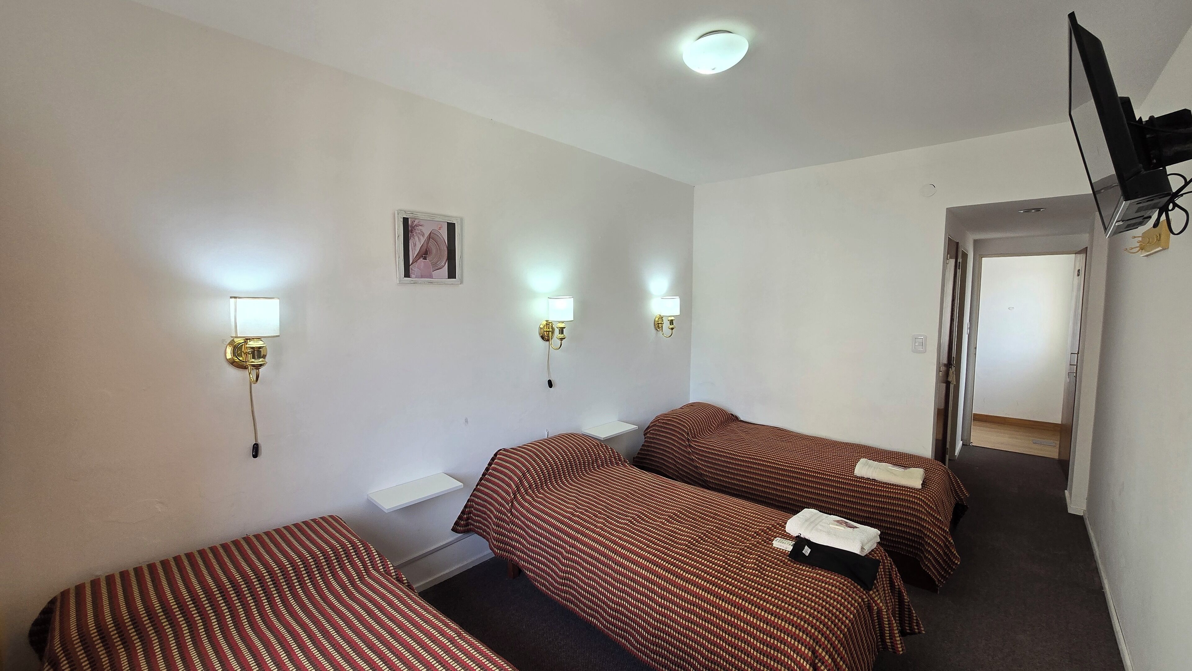 A hotel room with two single beds, a small bedside table, a wall-mounted TV, and two wall sconces.