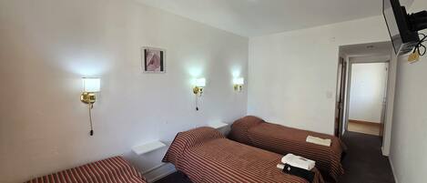 A hotel room with two single beds, a small bedside table, a wall-mounted TV, and two wall sconces.