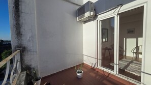 Un balcon agrémenté d’un pot de fleurs, une porte-fenêtre coulissante donnant sur un espace intérieur avec une table et des chaises, et un climatiseur fixé au mur.