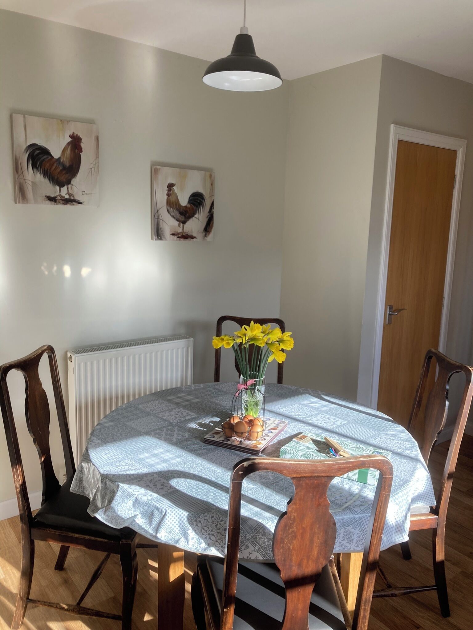 A dining area with a round table covered by a patterned tablecloth, surrounded by wooden chairs. A vase with yellow flowers is placed on the table, and there are two framed rooster paintings on the wall.
