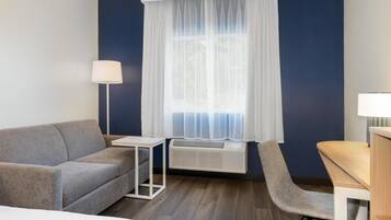 A hotel room with a grey sofa, a small table, a chair, a desk, and a window with sheer curtains.