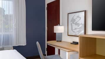A hotel room with a bed, a desk with a chair, a lamp, and a framed artwork on the wall.