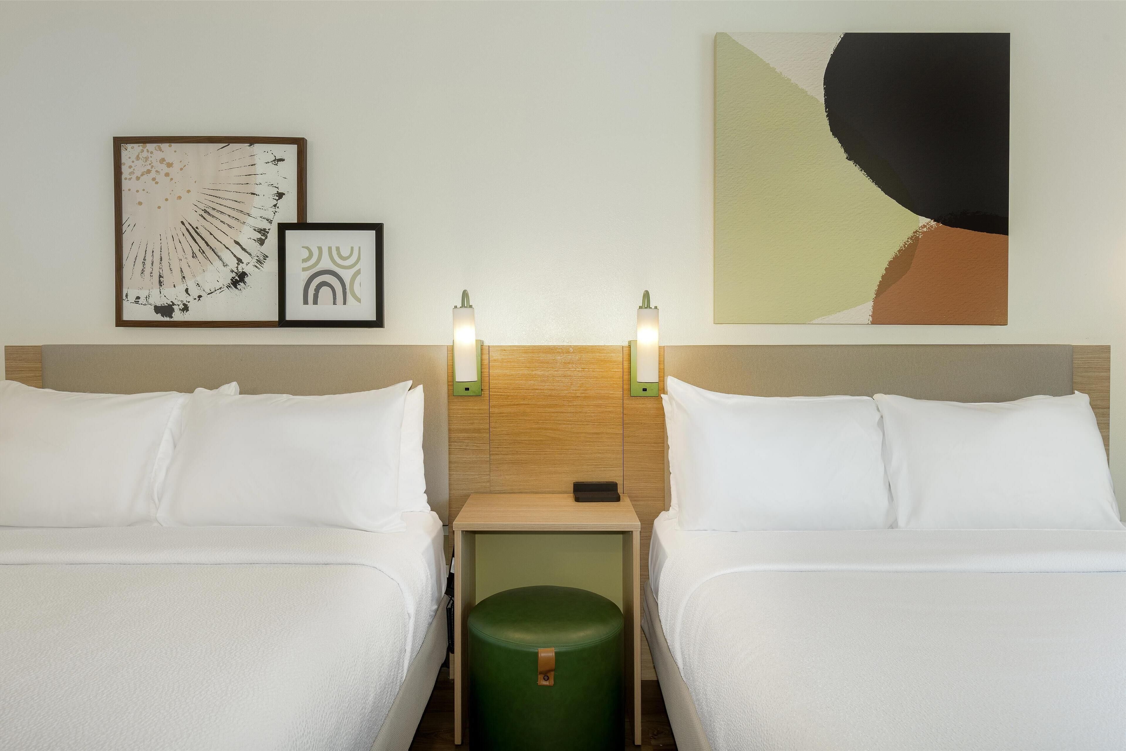 A hotel room with two beds, a green ottoman, wall art, and bedside lamps.
