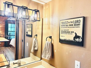 A bathroom with a mirror, sink, and towel rack. There is a framed quote on the wall and a window with curtains. - Entire Cabin in Branson, Missouri. Close to everything yet secluded for comfort. (Branson)