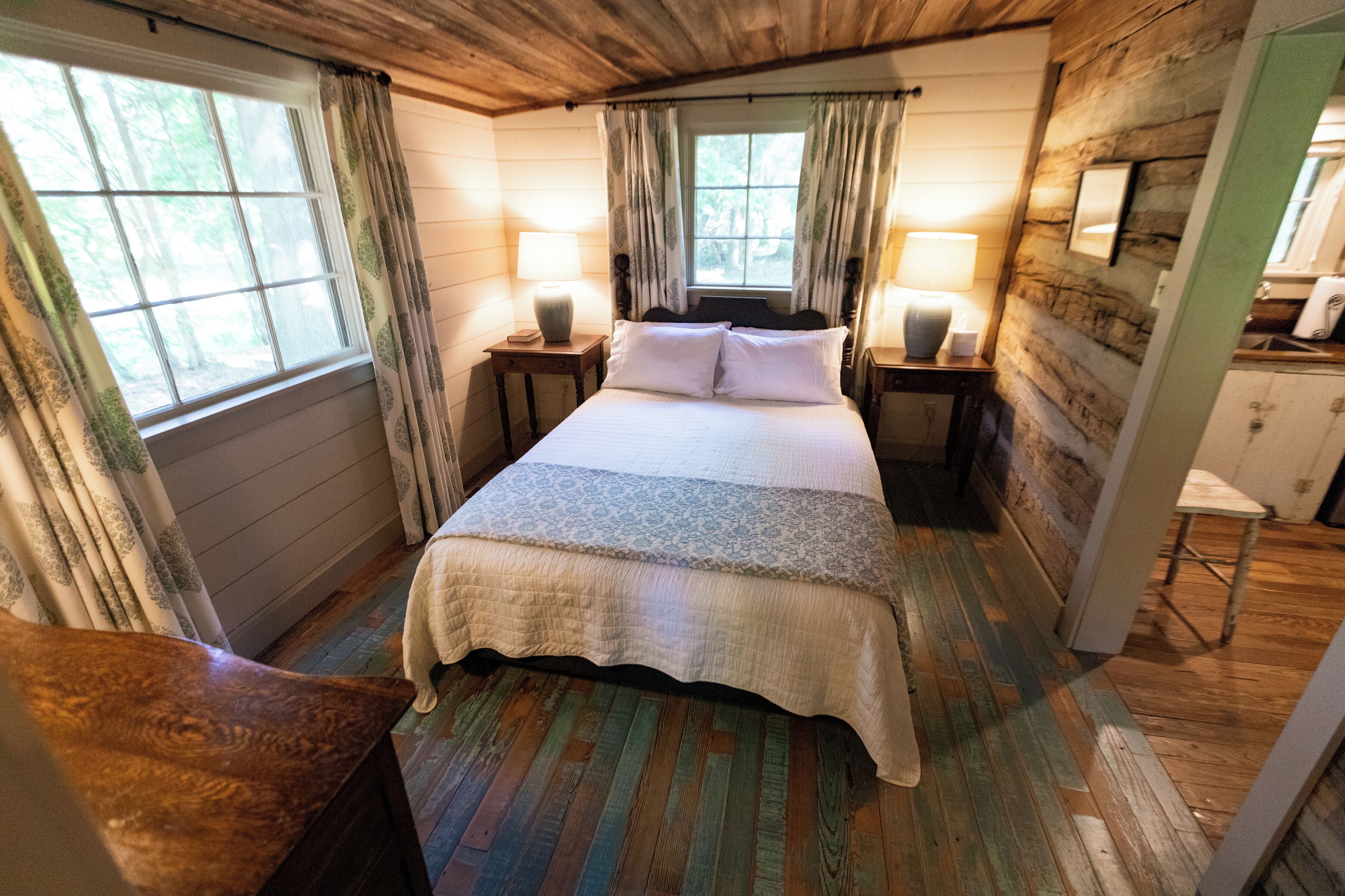 Ein Schlafzimmer in einer Holzhütte mit einem Bett, zwei Nachttischen mit Lampen, einer kleinen Holzkammer und einem Fenster mit Vorhängen.