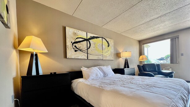 Une chambre d’hôtel avec un grand lit, deux tables de chevet avec des lampes, une chaise, un grand tableau abstrait au mur et une fenêtre donnant sur un paysage.