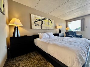 A hotel room with a large bed, two bedside tables with lamps, a chair, a large abstract painting on the wall, and a window providing a view. - Downtown CF Studio - Free Gated Parking! 521 (Cedar Falls)