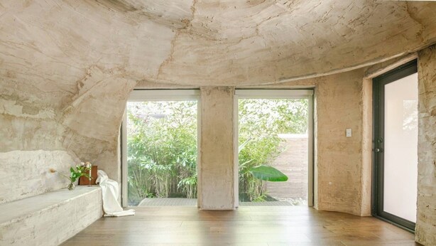 Il s’agit d’une pièce spacieuse avec un plafond haut et des murs en béton apparent. Deux grandes portes-fenêtres coulissantes donnent sur un jardin luxuriant. La pièce est dotée d’un plancher en bois et d’un banc le long du mur gauche.