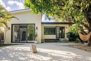 A modern house with a paved courtyard, large sliding glass doors, and a hammock under a tree. - Lumishi @_lumicollection (Miami)