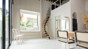 Un intérieur moderne avec un escalier, un coin repas avec des chaises et une grande fenêtre laissant entrer la lumière naturelle.