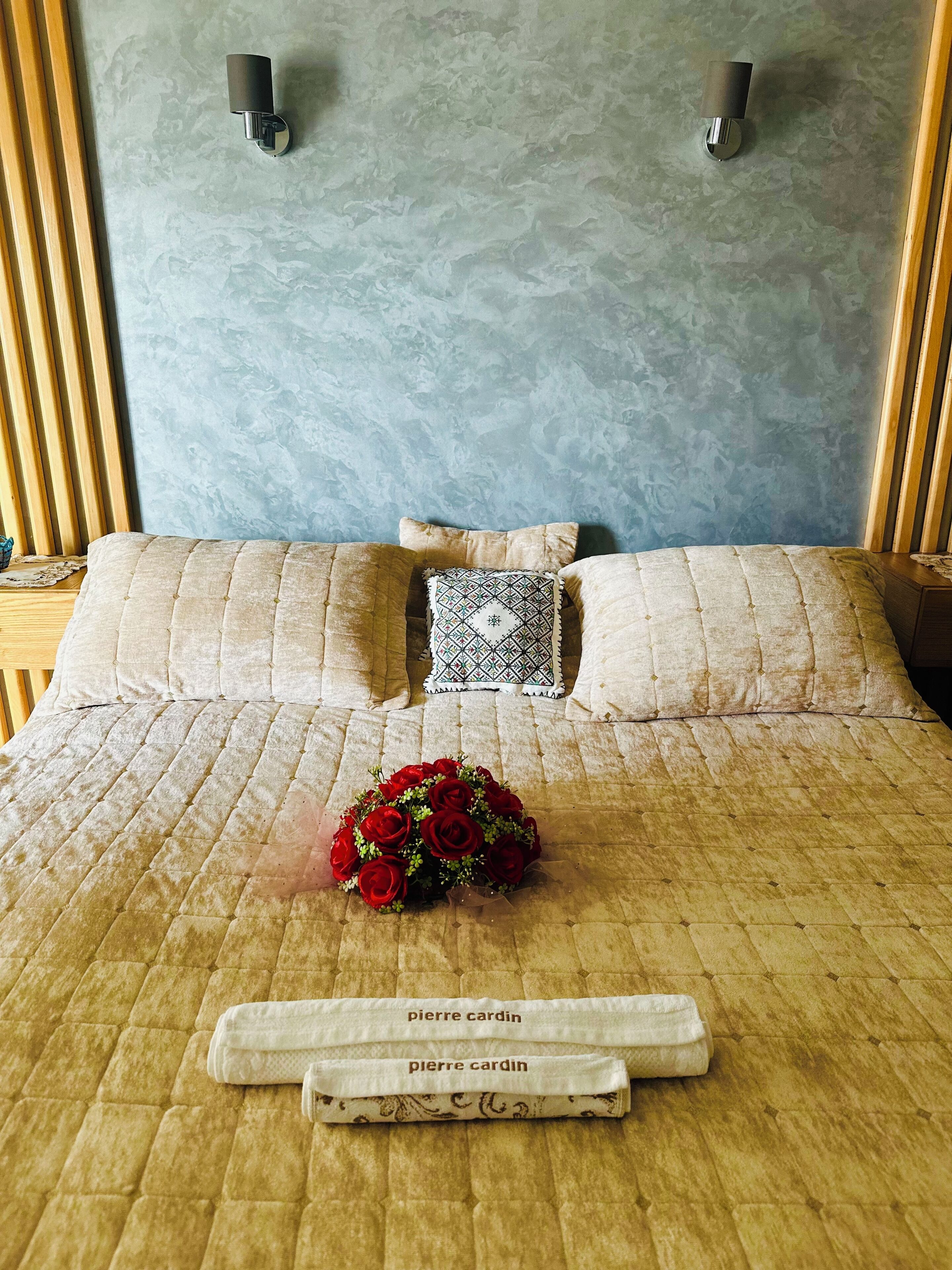 A neatly made bed with a quilted cover, a bouquet of red roses, and two folded towels labeled "pierre cardin" on a wooden floor.