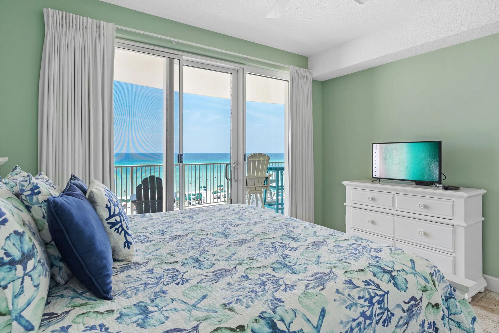 A bedroom with a bed, a TV on a dresser, and a view of the ocean.