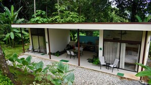 Un área de estar al aire libre con techo, con una mesa y sillas, rodeada de follaje y un camino de grava.