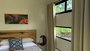Habitación de hotel con cama, un cuadro de colibrí, ventana con cortinas y cabecero de madera.