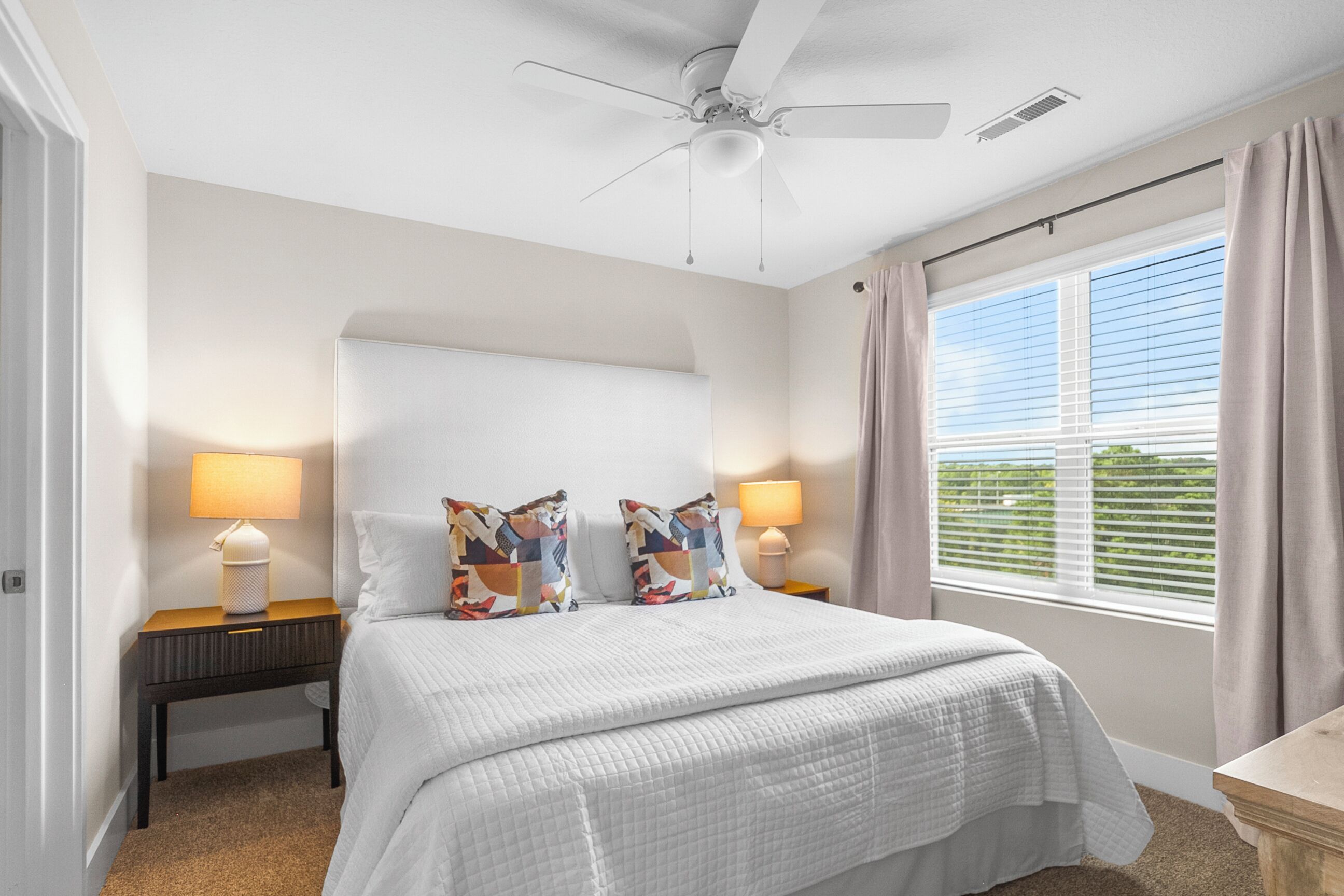 A bedroom with a bed, two bedside tables with lamps, a ceiling fan, a window with curtains, and a view of greenery outside.