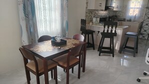 Zona pranzo con tavolo apparecchiato per quattro, circondato da sedie. Si intravede in secondo piano una cucina con elettrodomestici e mobili moderni.