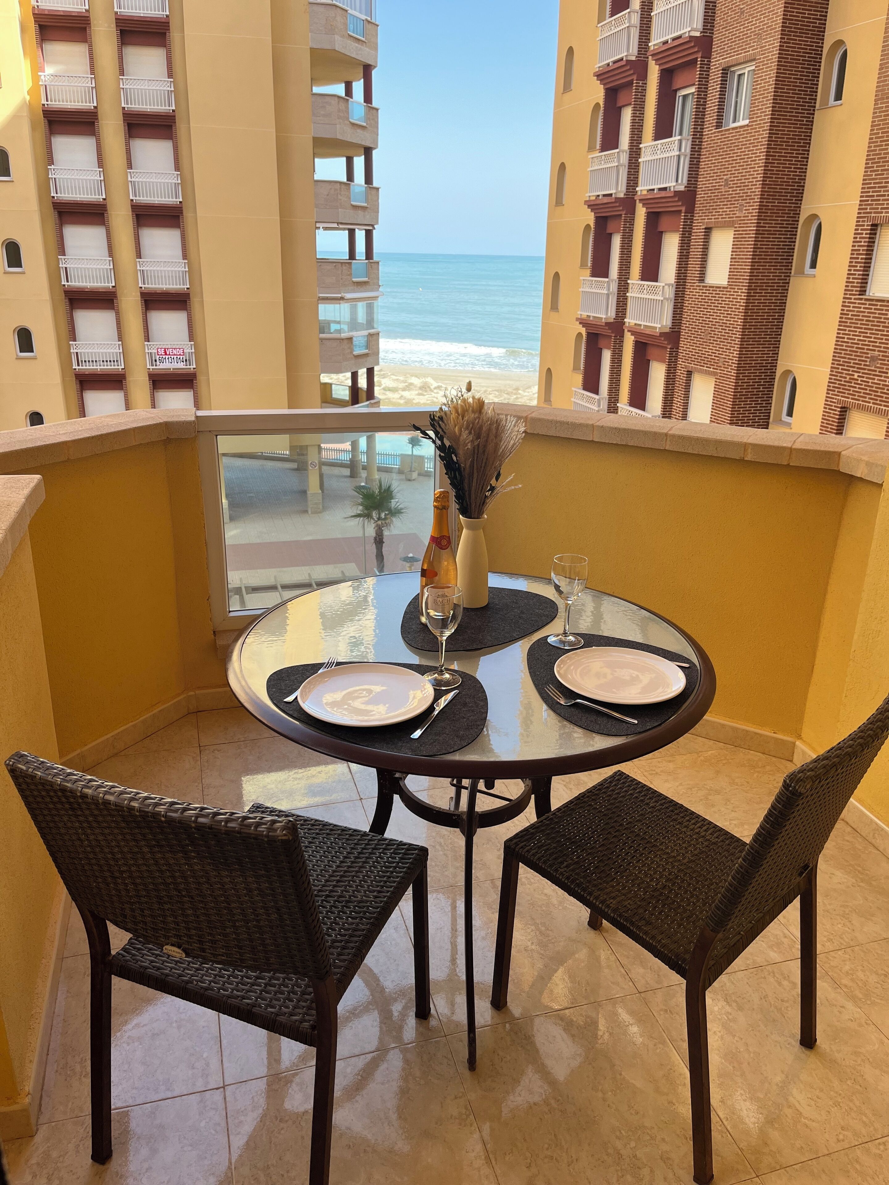 A balcony with a table set for two, overlooking a beach and high-rise buildings.