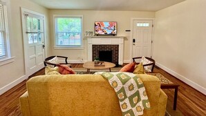 A living room with a fireplace, a television, and a yellow sofa. - Tea Olive Cottage - 2BR Near Augusta National, Downtown & Medical District (Augusta)