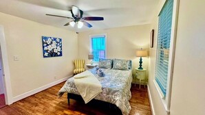 A bedroom with a bed, a ceiling fan, a window with blinds, a lamp, and a painting on the wall. - Tea Olive Cottage - 2BR Near Augusta National, Downtown & Medical District (Augusta)