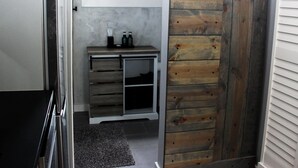 A bathroom with a wooden wall, a small vanity with a mirror, and a window above the mirror. - Hits | Catskills Views | Pet Friendly | Studio (Saugerties)