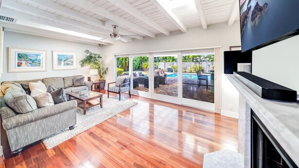 A spacious living room with a grey sofa, wooden coffee table, and a fireplace. Large windows offer a view of a backyard with outdoor furniture and a pool.
