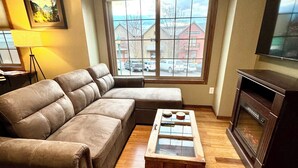 Ein Wohnzimmer mit Ecksofa, Glastisch, Kamin und einem großen Fenster mit Blick auf Gebäude und Autos draußen.