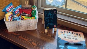 Ein Korb mit Snacks steht auf einer Theke neben einem Fenster mit Blick auf eine verschneite Straße. Ein Kreidetafel zeigt den Text: 'Gönnen Sie sich einen Snack'.