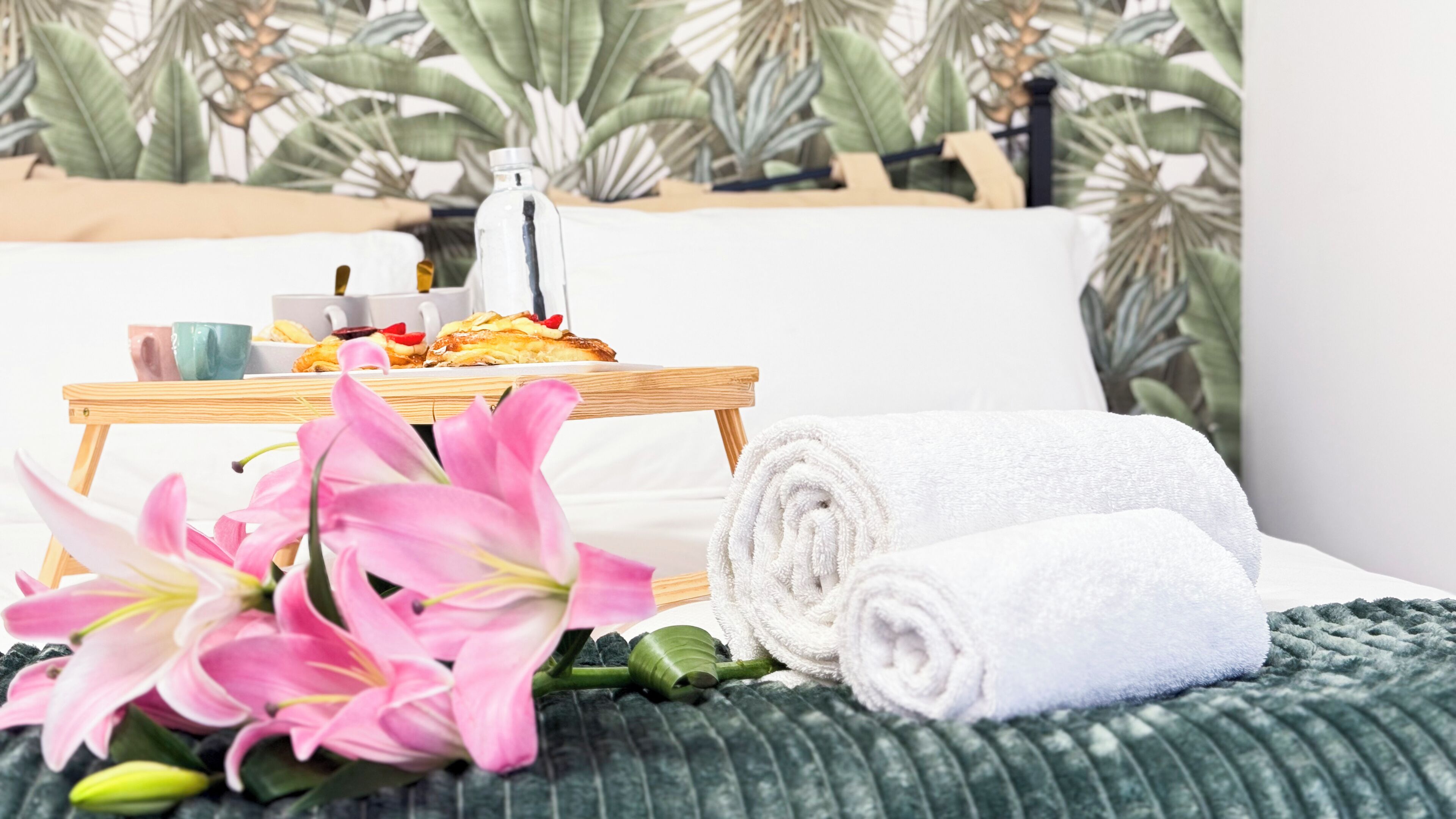 A neatly made bed with white towels, a tray with pastries, a bottle of water, and pink flowers on a green textured bedspread.