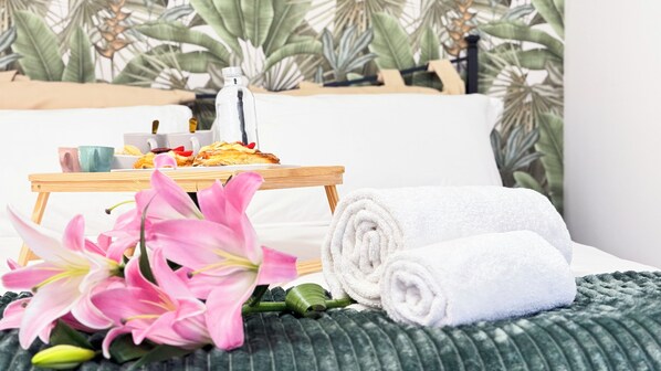 A neatly made bed with white towels, a tray with pastries, a bottle of water, and pink flowers on a green textured bedspread.