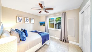 Une chambre à coucher avec un lit, un ventilateur de plafond, des œuvres d’art accrochées au mur et une fenêtre avec des rideaux.