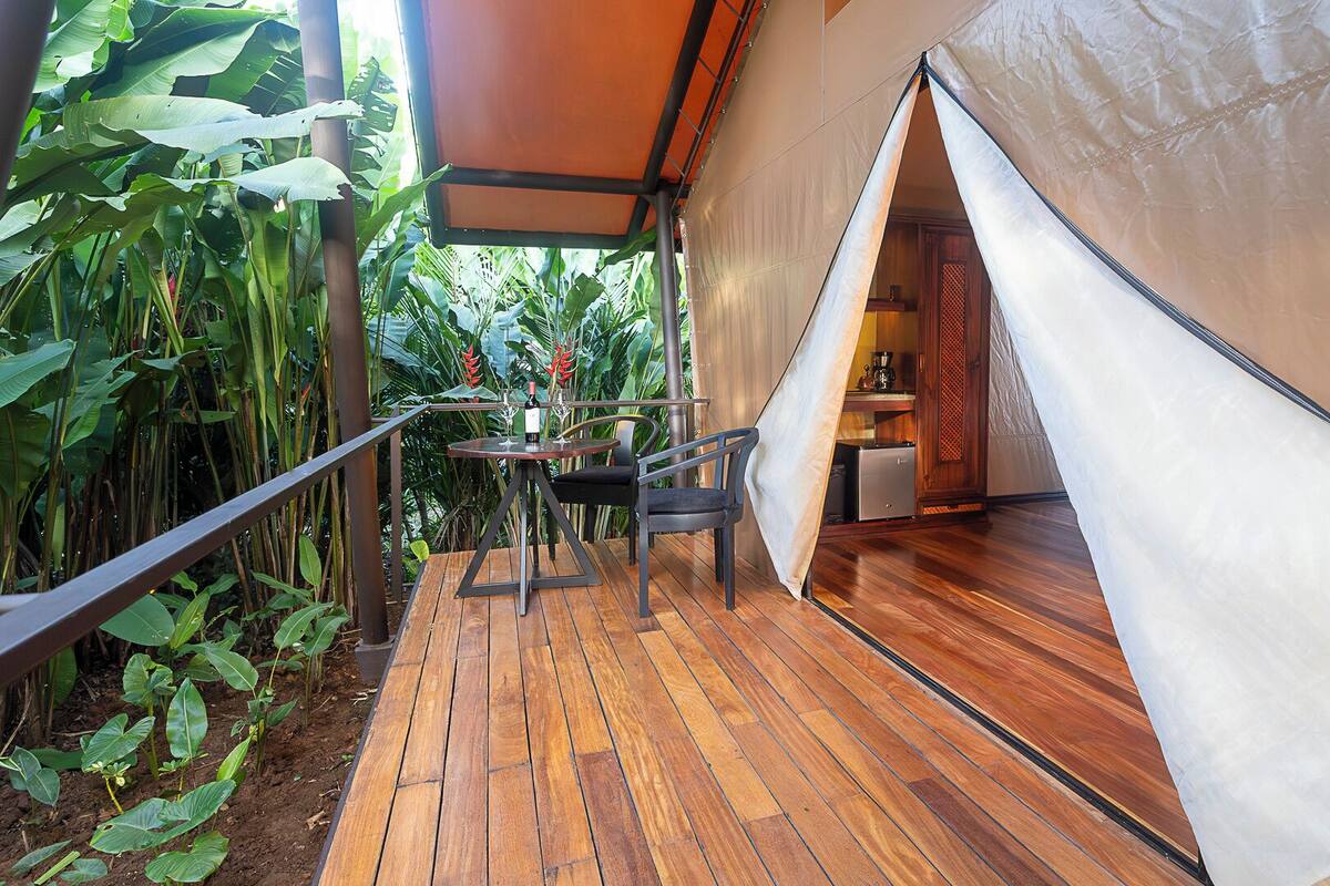 A tented accommodation with a wooden deck, a table with chairs, and a view of lush greenery.
