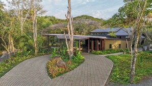 A modern house with a covered patio, surrounded by lush greenery and a paved walkway.