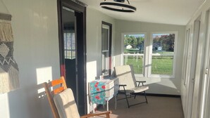 Une pièce avec un fauteuil à bascule, une petite table sur laquelle est posée une courtepointe, et une vue sur une pelouse verdoyante par la fenêtre.