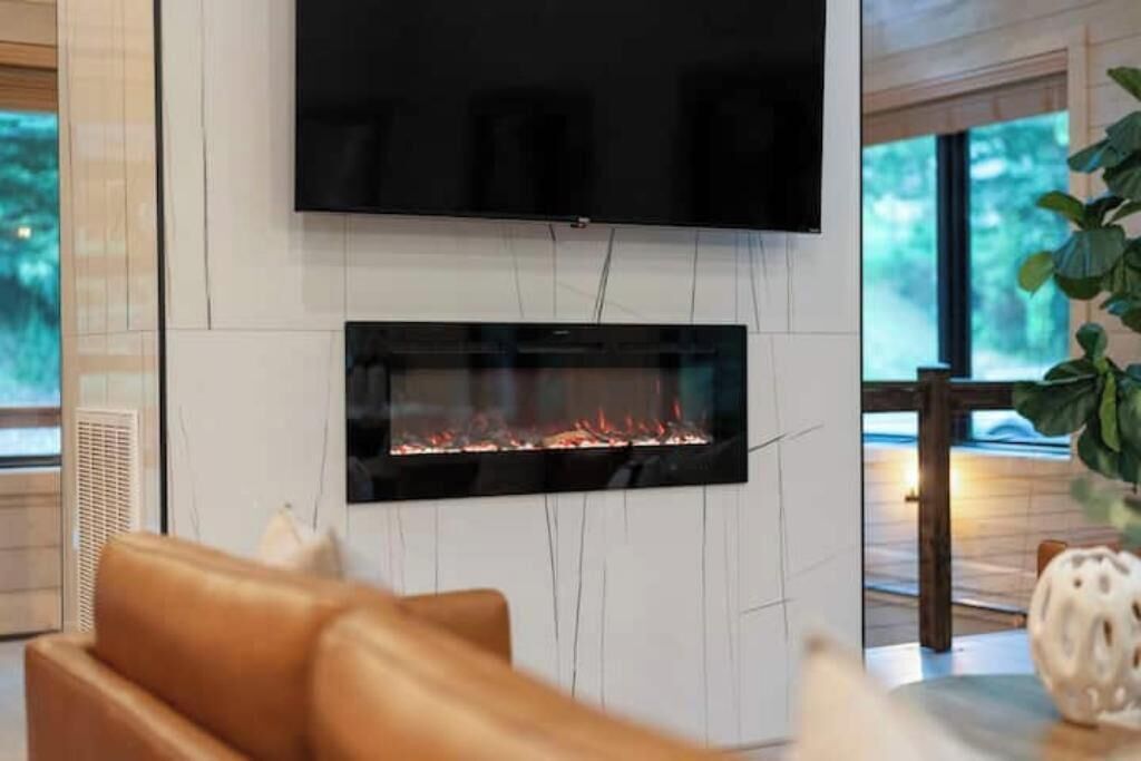 A modern living room with a flat-screen TV mounted above a glass fireplace, a leather sofa, and a decorative plant.