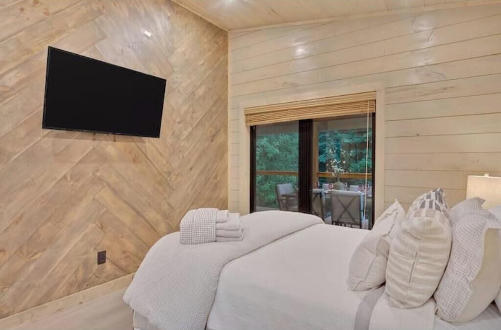 A modern bedroom with a herringbone-patterned wall, a flat-screen TV mounted above a bed, and a large window offering a view of outdoor furniture.