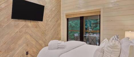 A modern bedroom with a herringbone-patterned wall, a flat-screen TV mounted above a bed, and a large window offering a view of outdoor furniture.