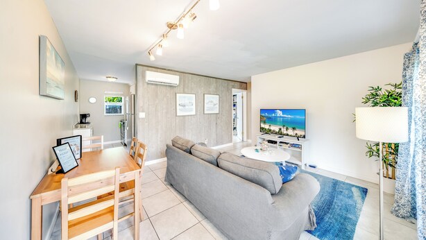 A modern living room with a grey sofa, wooden dining table, and chairs. There is a TV on the wall, an air conditioning unit, and a small kitchen area in the background. - Beach Unit! Walk To Beach & Restaurants / Shops! (Pompano Beach)