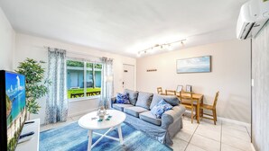 A living room with a sofa, a dining table, and chairs. There is a TV on a stand, a plant, and a window with curtains. - Beach Unit! Walk To Beach & Restaurants / Shops! (Pompano Beach)