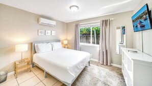 A hotel room with a bed, bedside table, lamp, air conditioning unit, window with curtains, and a TV. - Beach Unit! Walk To Beach & Restaurants / Shops! (Pompano Beach)