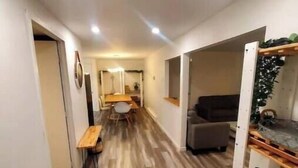 Un pasillo con piso de madera, una zona de comedor con mesa y sillas, un sofá y un mueble de estanterías con objetos.