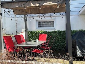 Une terrasse extérieure avec une table et des chaises rouges, des guirlandes lumineuses suspendues au-dessus, et une fenêtre donnant sur un bosquet.