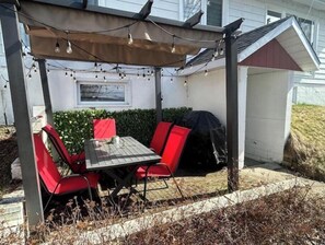 Une terrasse extérieure avec une table et des chaises rouges, une pergola illuminée de guirlandes lumineuses et un espace barbecue abrité.