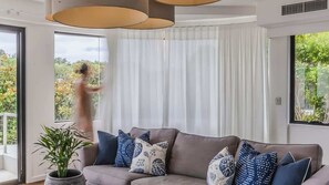 A modern living room with a sofa, patterned cushions, a potted plant, and large windows with curtains.
